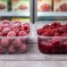 Two clear plastic containers on a wooden surface: frosted raspberries on the left and raspberry jam on the right, with a grocery freezer in the background.