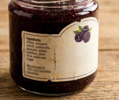 Jar of blackberry jam on a wooden table with a beige label and a blackberry illustration on the side label.