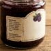 Jar of blackberry jam on a wooden table with a beige label and a blackberry illustration on the side label.