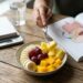 Person studying at a wooden table with an open anatomy book, a glass of water, a notebook, and a bowl of fresh fruit (banana slices, grapes, mango, pineapple).