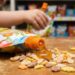 Child's hand tilts a colorful snack pouch to spill fruit-shaped gummies onto a wooden table.