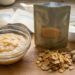 A DIY baby-food prep scene on a wooden board: a glass bowl of smooth purée, a metallic pouch with a window showing its contents, dried cereal-like flakes, small white berries, and a dropper bottle of oil beside a white bowl.