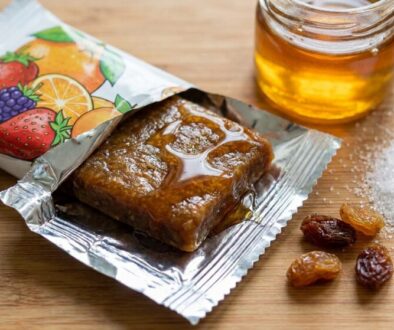 Open fruit snack bar on a foil wrapper with a drizzle of syrup, next to a jar of honey, sugar granules, and raisins on a wooden surface