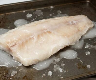 Raw white fish fillet on a baking sheet with scattered ice and juices around it, ready to cook.