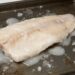Raw white fish fillet on a baking sheet with scattered ice and juices around it, ready to cook.
