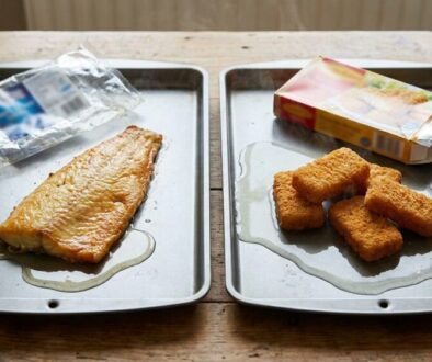 Two baking sheets on a wooden table—left with a raw fish fillet and drippings, right with breaded nuggets and an open box nearby.