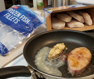 Frying pan on a stove with a thick fish fillet cooking in butter and a smaller piece beside it; a bag of frozen fish fillets and an open box of bread or biscuits on the counter nearby in a kitchen.