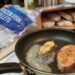 Frying pan on a stove with a thick fish fillet cooking in butter and a smaller piece beside it; a bag of frozen fish fillets and an open box of bread or biscuits on the counter nearby in a kitchen.