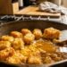 Cast-iron skillet on a stove with golden fried chicken pieces simmering in a brown sauce; a spatula rests inside the pan.