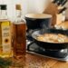 Skillet on a gas stove sizzling with oil, nearby are potatoes, a rosemary sprig, and two oil bottles on a wooden counter.
