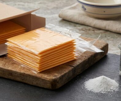Stack of individually wrapped orange cheese slices on a wooden cutting board next to an open cardboard box.