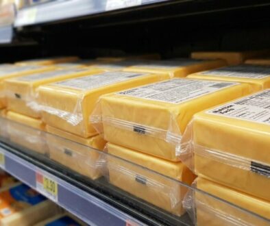 Blocks of yellow cheese wrapped in plastic on a supermarket shelf, close view of packaging and price tags nearby.
