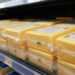 Blocks of yellow cheese wrapped in plastic on a supermarket shelf, close view of packaging and price tags nearby.