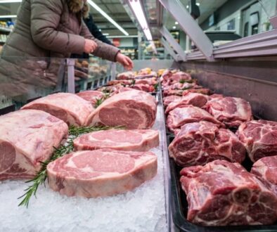 Beef ribeye steaks laid on crushed ice at a supermarket meat counter, with a shopper reaching for meat in the background and rosemary garnish nearby.