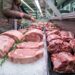 Beef ribeye steaks laid on crushed ice at a supermarket meat counter, with a shopper reaching for meat in the background and rosemary garnish nearby.