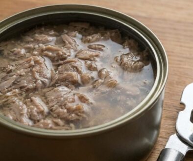 Open can of tuna with shredded tuna meat in brine on a wooden surface next to a metal can opener.