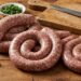 Coiled raw sausages on a wooden countertop with garlic, herbs, and a knife nearby on a cutting board.