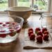 Strawberries being rinsed in a glass bowl of water on a kitchen counter, with drying berries on napkins nearby and a jar in the background.