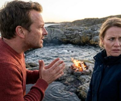Man in a red sweater speaks with hands clasped as a worried woman in a dark jacket stands by a small campfire on rocky coastline at sunset.