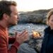 Man in a red sweater speaks with hands clasped as a worried woman in a dark jacket stands by a small campfire on rocky coastline at sunset.