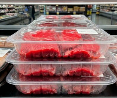 Stack of clear plastic meat trays filled with red beef in a supermarket meat section, condensation on the lids and price labels visible in the background.