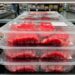Stack of clear plastic meat trays filled with red beef in a supermarket meat section, condensation on the lids and price labels visible in the background.
