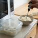 In a kitchen, a hand inserts a digital thermometer into a bowl of cooked rice on the counter, with a glass container of rice nearby and a microwave in view nearby.