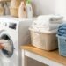 Person loading a front-loading washing machine with a white towel in a laundry room, surrounded by detergent bottles and baskets.