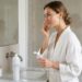 Woman in a white robe applies skincare to her face at a bathroom sink vanity.