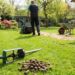 People gardening in a backyard with a soil aerator, rake, wheelbarrow, and a pile of soil cores on the lawn.