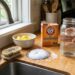 Hand holding a glass jar of water beside a box of baking soda on a wooden kitchen counter. A mound of baking soda sits on a metal dish nearby, with a brush and lemons in the background.
