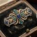 Ornate vintage brooch with blue, green, and white stones resting in an old velvet-lined wooden jewelry box, with a magnifying glass beside it.