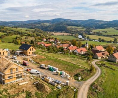 Aerial view of a construction site with wooden houses under building, set on a hillside beyond a rural village and green fields laid out with rolling hills in the distance.