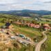 Aerial view of a construction site with wooden houses under building, set on a hillside beyond a rural village and green fields laid out with rolling hills in the distance.