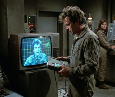 Man in a dusty control room uses a handheld console while a blue-tinted screen shows a man on a grid background.