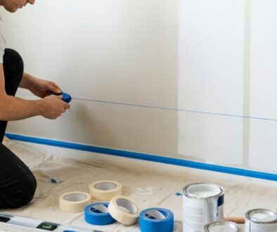 Person kneels on a drop cloth, tightening a blue string to mark a straight line on a white wall for painting prep. Nearby are masking tape rolls and paint cans.