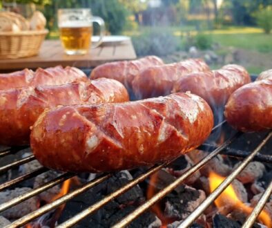 Sausages grilling on a barbecue over hot coals with a basket of bread and a beer mug in the background.