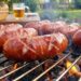 Sausages grilling on a barbecue over hot coals with a basket of bread and a beer mug in the background.