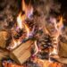 A cozy fireplace with stacked logs and pinecones burning, flames licking the wood and smoke curling upward, brick surround visible