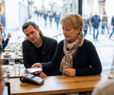 Woman at a cafe counter paying with a card on a portable reader while companions sit at the table and pedestrians pass outside the windowed street entrance, daytime scene.