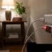 Close-up of a wall outlet with two USB power adapters plugged in; a small device glows red, while a lamp-lit living room with a side table and plant is in the blurred background.