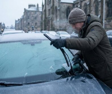 Man in a brown winter coat and knit hat scrapes ice off a car windshield on a snowy street corner.