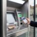 Person using a smartphone at an outdoor ATM, with cash visible in the tray and a card reader nearby.