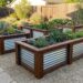 Raised wooden planters with corrugated metal sides filled with succulents and drought-tolerant plants in a gravel backyard. They form a tidy garden grid around a stone path.