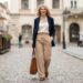 Woman in a navy blazer and beige trousers walks down a cobblestone street, carrying a brown tote.