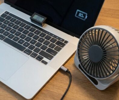 Laptop on a wooden desk with a small USB-powered fan resting on the top edge of the keyboard area; a larger portable desk fan sits to the right, plugged into the laptop. The screen shows an 80% battery icon.