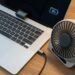 Laptop on a wooden desk with a small USB-powered fan resting on the top edge of the keyboard area; a larger portable desk fan sits to the right, plugged into the laptop. The screen shows an 80% battery icon.