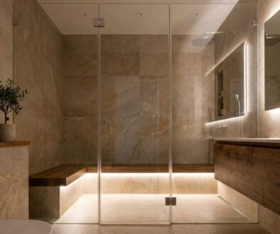 Modern beige-tiled bathroom with a glass walk-in shower, wooden vanity, a built-in bench, towels, and a potted plant on the counter.