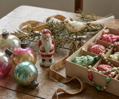 Assorted vintage Christmas ornaments on a rustic wooden table, including a Santa figure, glass baubles, a metal bird, and a bead garland in a box.