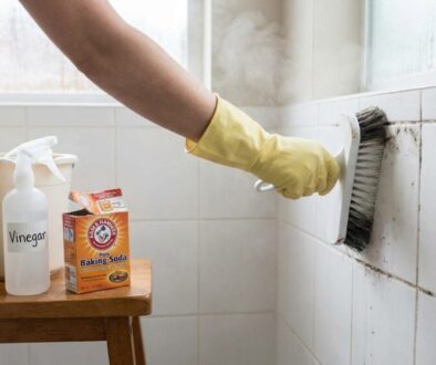 Person in yellow cleaning gloves scrubs dirty tile grout with a brush, while cleaning supplies (vinegar, baking soda, sponge) sit on a wooden stool nearby.